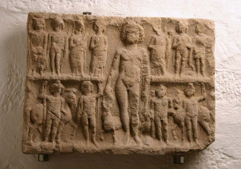 A full-color photo of the badly weathered brown sandstone landscape-oriented relief mounted on a wall. There are two rows of figures: seven human figures on the top, and four below. Mercury stands in the middle of Them at the full height of both rows. A cloak is clasped over His shoulder. The caduceus is in His left hand, perhaps the bag of money in His right, below which is the ram or goat. The figures paired on the lower row (two on each side of Mercury) each have a horse. See footnote \#5 for further detail.
